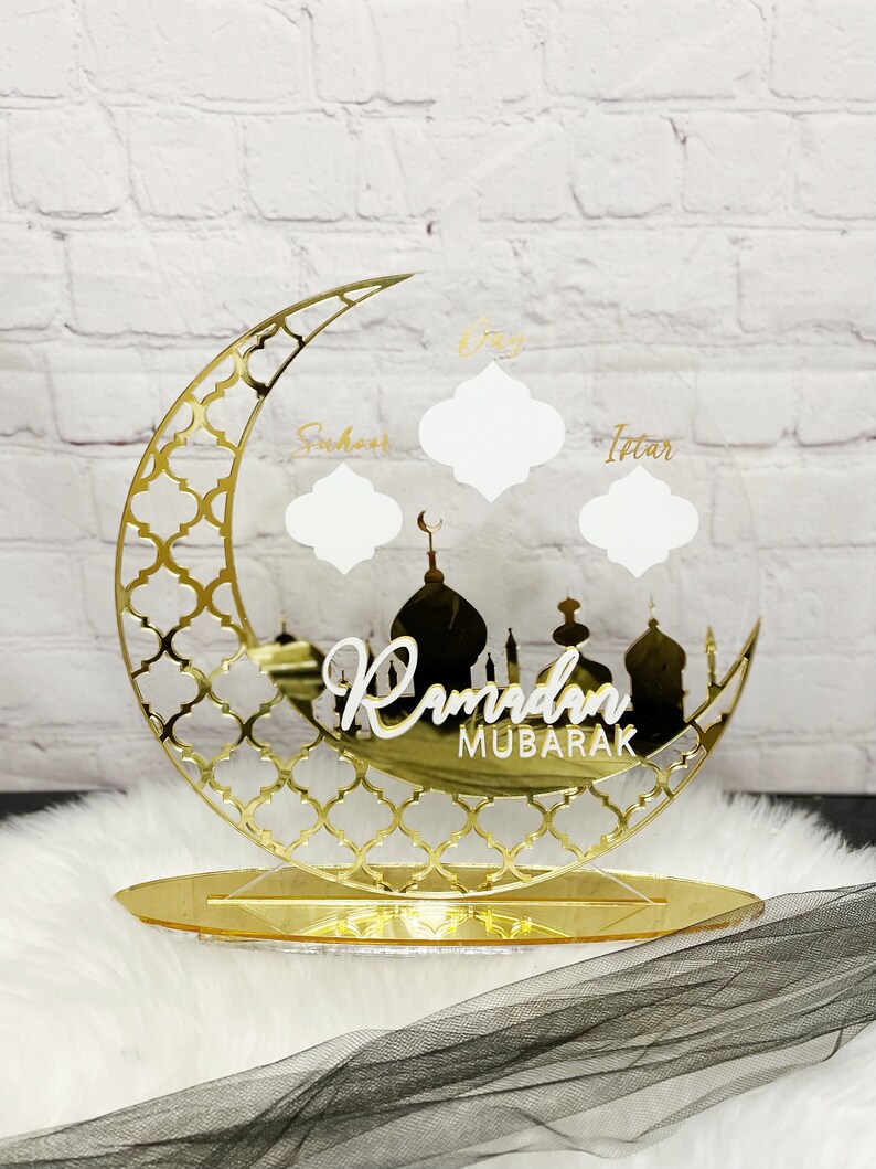 May include: Gold Ramadan Mubarak sign with a crescent moon and mosque silhouette. The sign has three blank spaces for writing in the names of the days of Ramadan: Suhoor, Iftar, and Day.