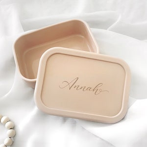 Pode incluir: Um recipiente retangular bege com uma tampa a condizer. A tampa apresenta o nome "Annah" em elegante escrita cursiva. O recipiente est&aacute; aberto, revelando o seu interior. O fundo &eacute; um tecido branco e sedoso.