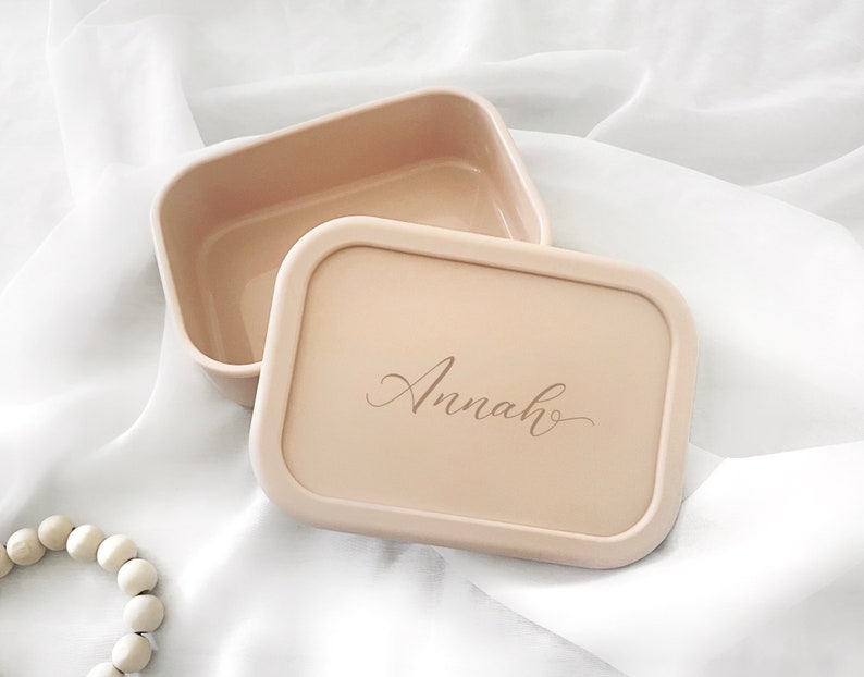 Pode incluir: Um recipiente retangular bege com uma tampa a condizer. A tampa apresenta o nome "Annah" em elegante escrita cursiva. O recipiente est&aacute; aberto, revelando o seu interior. O fundo &eacute; um tecido branco e sedoso.