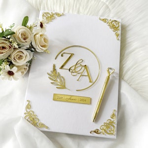 Puede incluir: Libro de visitas de boda blanco con detalles dorados. La portada presenta un círculo dorado con las iniciales "Z & A" y un diseño de hoja dorada. La fecha "21 de junio de 2024" está escrita en una placa dorada debajo del círculo.