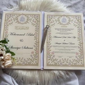 May include: Open wedding invitation book with floral border and calligraphy. The left page reads "Mohemmed-Bilal & Shaniya Sultana". The right page has similar design with text. A jeweled pen rests between the pages.
