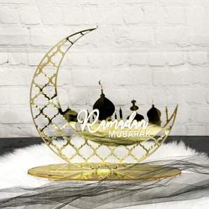 May include: A gold mirrored crescent moon with a decorative pattern and a silhouette of a mosque with the text "Ramadan Mubarak" in white lettering. The moon is on a gold stand.