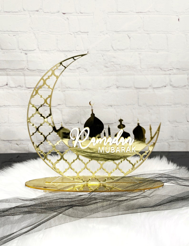 May include: A gold mirrored crescent moon with a decorative pattern and a silhouette of a mosque with the text "Ramadan Mubarak" in white lettering. The moon is on a gold stand.