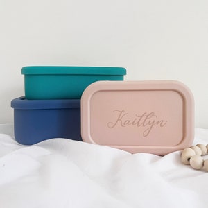 Pode incluir: Tr&ecirc;s recipientes retangulares para alimentos em verde-azulado, azul marinho e rosa. O recipiente rosa tem o nome "Kaitlyn" gravado na tampa. Os recipientes est&atilde;o empilhados.