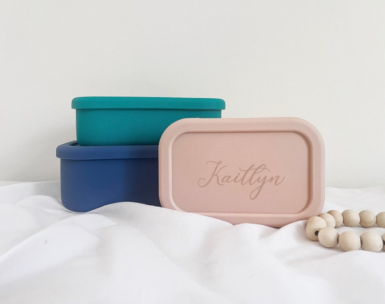 Pode incluir: Tr&ecirc;s recipientes retangulares para alimentos em verde-azulado, azul marinho e rosa. O recipiente rosa tem o nome "Kaitlyn" gravado na tampa. Os recipientes est&atilde;o empilhados.