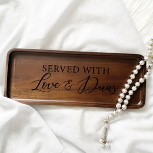 May include: A rectangular wooden tray with the words "SERVED WITH Love & Duas" engraved in black script. A string of white beaded prayer beads rests beside the tray on a white fabric surface.