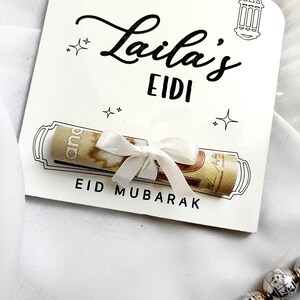 May include: A white card with black text that reads "Laila's Eidi Eid Mubarak". A rolled up bill of money is tied with a white ribbon and placed inside a drawn scroll on the card.