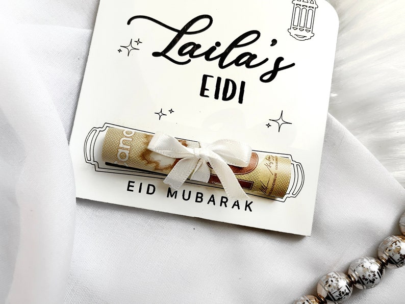 May include: A white card with black text that reads "Laila's Eidi Eid Mubarak". A rolled up bill of money is tied with a white ribbon and placed inside a drawn scroll on the card.