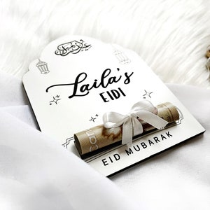 May include: White wooden sign with black calligraphy that reads "Laila's Eidi Eid Mubarak". A rolled up bill of money is tied with a white ribbon and placed on the sign.