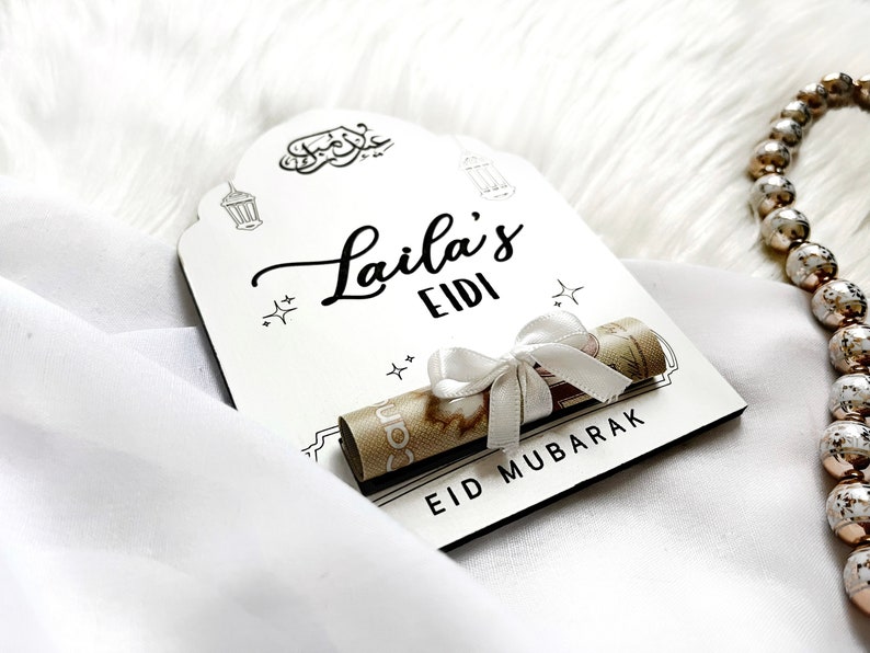 May include: White wooden sign with black calligraphy that reads "Laila's Eidi Eid Mubarak". A rolled up bill of money is tied with a white ribbon and placed on the sign.