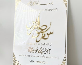 May include: A white wedding sign with gold accents and calligraphy. The sign reads "WELCOME TO OUR WEDDING" and features the names "SABREEN & SARMAD". Decorative gold corner designs and text in Arabic are also present.