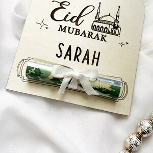 Puede incluir: Un letrero de madera con el texto "Eid Mubarak Sarah" y un billete enrollado atado con una cinta blanca.