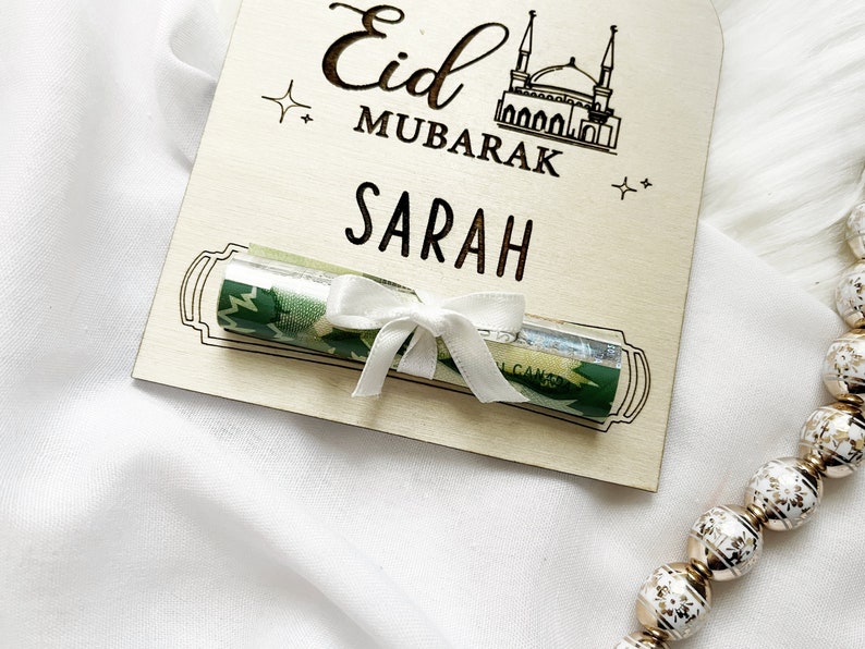 Puede incluir: Un letrero de madera con el texto "Eid Mubarak Sarah" y un billete enrollado atado con una cinta blanca.