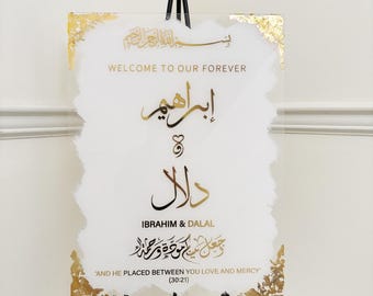 May include: A white acrylic sign with gold accents and calligraphy. The sign reads "Welcome to our forever" in English and Arabic. The names "Ibrahim & Dalal" are written in Arabic calligraphy. The sign also includes a verse from the Quran in Arabic calligraphy, which translates to "And He placed between you love and mercy." (30:21)