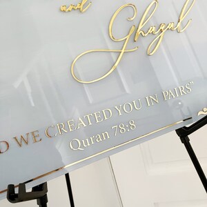 May include: A white sign with gold lettering that reads "And Ghazal" and "And we created you in pairs" Quran 78:8. The sign is on a black easel.
