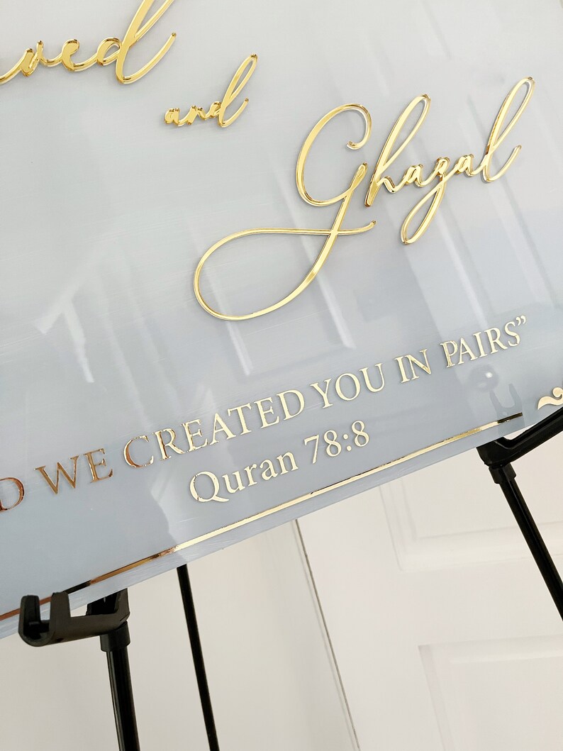 May include: A white sign with gold lettering that reads "And Ghazal" and "And we created you in pairs" Quran 78:8. The sign is on a black easel.