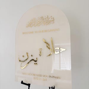 Puede incluir: Un letrero de acr&iacute;lico blanco con letras doradas. El letrero presenta la frase &aacute;rabe "Bismillah ar-Rahman ar-Rahim" que se traduce como "En el nombre de Al&aacute;, el M&aacute;s Misericordioso, el M&aacute;s Compasivo." El letrero tambi&eacute;n incluye el texto "Bienvenido a nuestro comienzo" y "Y te creamos en parejas" con una referencia al vers&iacute;culo del Cor&aacute;n.