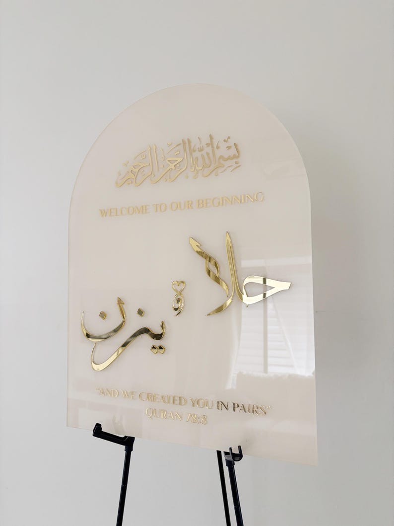 Puede incluir: Un letrero de acr&iacute;lico blanco con letras doradas. El letrero presenta la frase &aacute;rabe "Bismillah ar-Rahman ar-Rahim" que se traduce como "En el nombre de Al&aacute;, el M&aacute;s Misericordioso, el M&aacute;s Compasivo." El letrero tambi&eacute;n incluye el texto "Bienvenido a nuestro comienzo" y "Y te creamos en parejas" con una referencia al vers&iacute;culo del Cor&aacute;n.
