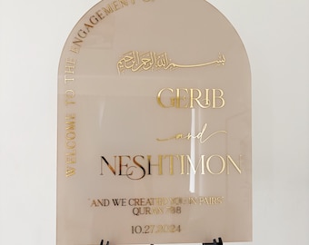May include: A beige acrylic welcome sign with gold lettering announcing the engagement of Gerib and Neshtimon. The sign includes an Arabic inscription and the date 10.27.2024.