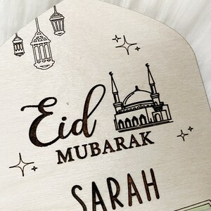 Puede incluir: Un letrero de madera con el texto "Eid Mubarak Sarah" en una fuente cursiva negra. El letrero tambi&eacute;n presenta una mezquita y estrellas.
