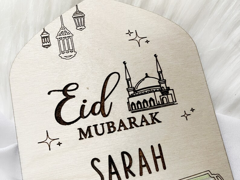 Puede incluir: Un letrero de madera con el texto "Eid Mubarak Sarah" en una fuente cursiva negra. El letrero tambi&eacute;n presenta una mezquita y estrellas.