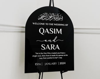 May include: Black acrylic wedding sign with an arch shape. The sign features the Arabic phrase "Bismillah ar-Rahman ar-Rahim" at the top, followed by "Welcome to the Wedding of Qasim and Sara". The sign also includes a quote from the Quran: "He is the One Who created you from a single soul, then from it made its spouse so he may find comfort in her" (7:189). The date of the wedding is listed below the quote: "13th | January | 2024".