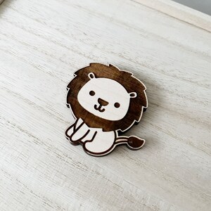 May include: A wooden lion cutout with a brown mane and white face. The lion is sitting with its paws tucked under its body and has a small tail.