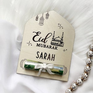 Puede incluir: Una tarjeta de madera con un dise&ntilde;o de mezquita y linternas. La tarjeta dice "Eid Mubarak" y "Sarah". Un billete enrollado est&aacute; atado con una cinta blanca y colocado en la tarjeta.