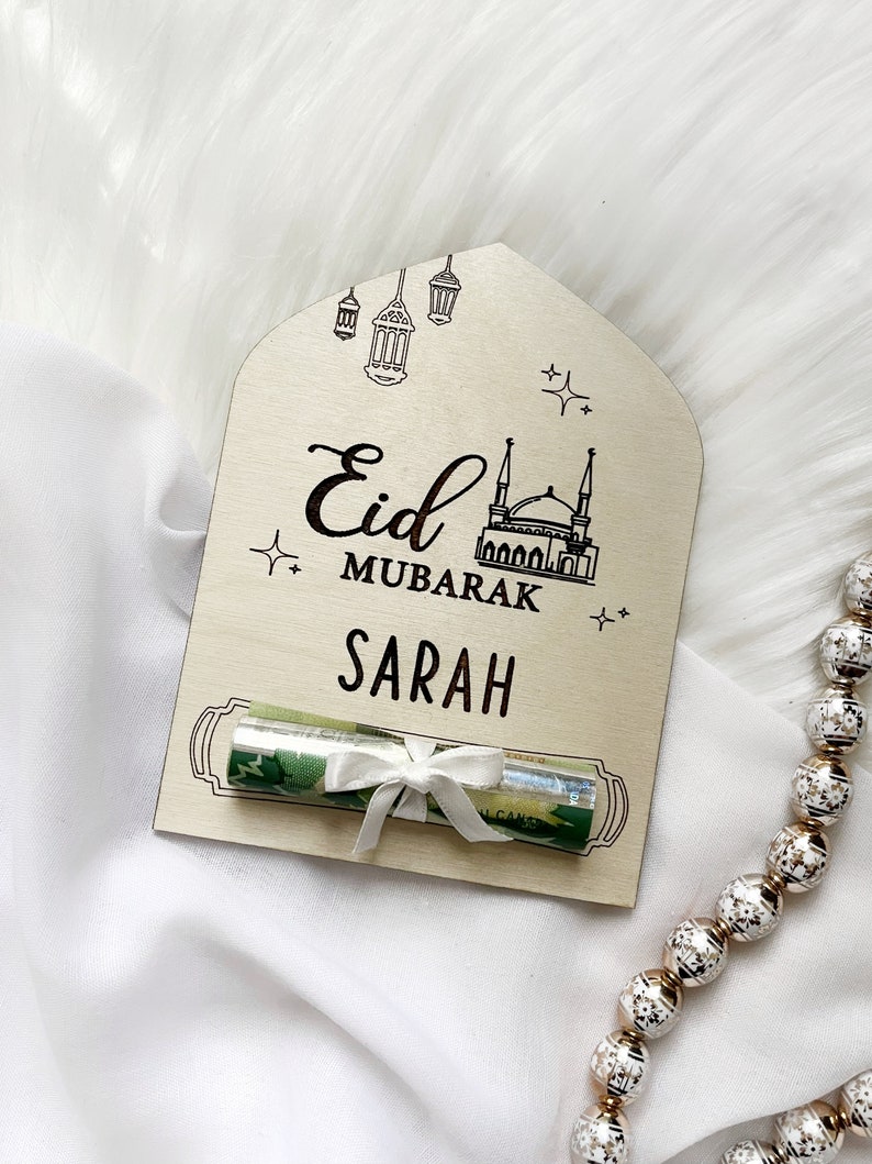 Puede incluir: Una tarjeta de madera con un dise&ntilde;o de mezquita y linternas. La tarjeta dice "Eid Mubarak" y "Sarah". Un billete enrollado est&aacute; atado con una cinta blanca y colocado en la tarjeta.