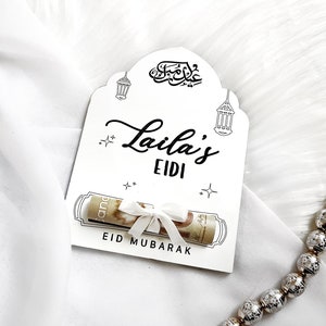 May include: White card with black calligraphy that says "Eid Mubarak" and "Laila's Eidi". A rolled up bill of money is tied with a white ribbon and placed inside the card.