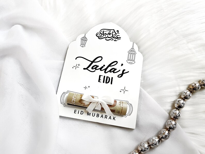 May include: White card with black calligraphy that says "Eid Mubarak" and "Laila's Eidi". A rolled up bill of money is tied with a white ribbon and placed inside the card.