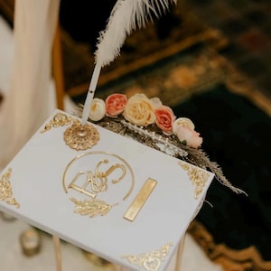 May include: White guest book with gold accents and a gold monogram of "E&S" on the front. A white feather pen is placed on top of the book. The book is decorated with a floral arrangement of peach and cream flowers.