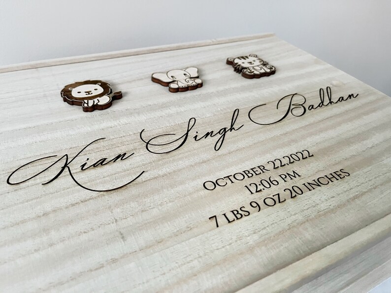 May include: A wooden keepsake box with a light wood finish. The box is engraved with the name "Kian Singh Badhan", the date "October 22, 2022", the time "12:06 PM", and the weight and length "7 LBS 9 OZ 20 INCHES". The box is decorated with three wooden animal cutouts: a lion, an elephant, and a tiger.