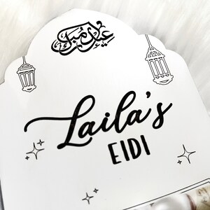 May include: White wooden sign with black calligraphy that says "Laila's Eidi" with two black and white illustrations of lanterns and stars.