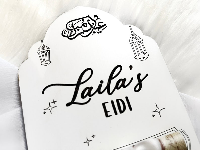 May include: White wooden sign with black calligraphy that says "Laila's Eidi" with two black and white illustrations of lanterns and stars.