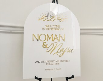 May include: White arch-shaped sign with gold lettering announcing the wedding of Noman & Majra. The sign includes an Arabic phrase, the date November 17, 2023, and the quote "And we created you in pairs" from the Quran 78:8.