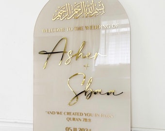 May include: A wedding sign with an arched top. The sign is a light beige color with gold lettering. The text includes Arabic script, "WELCOME TO THE WEDDING OF Asher + Shan", a quote, and the date 05.11.2024.