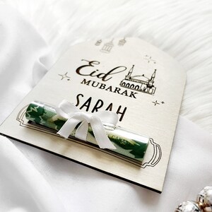Puede incluir: Una tarjeta de madera con el texto "Eid Mubarak Sarah" y una tarjeta de regalo enrollada verde y blanca con una cinta blanca. La tarjeta tiene un dibujo de una mezquita y estrellas.