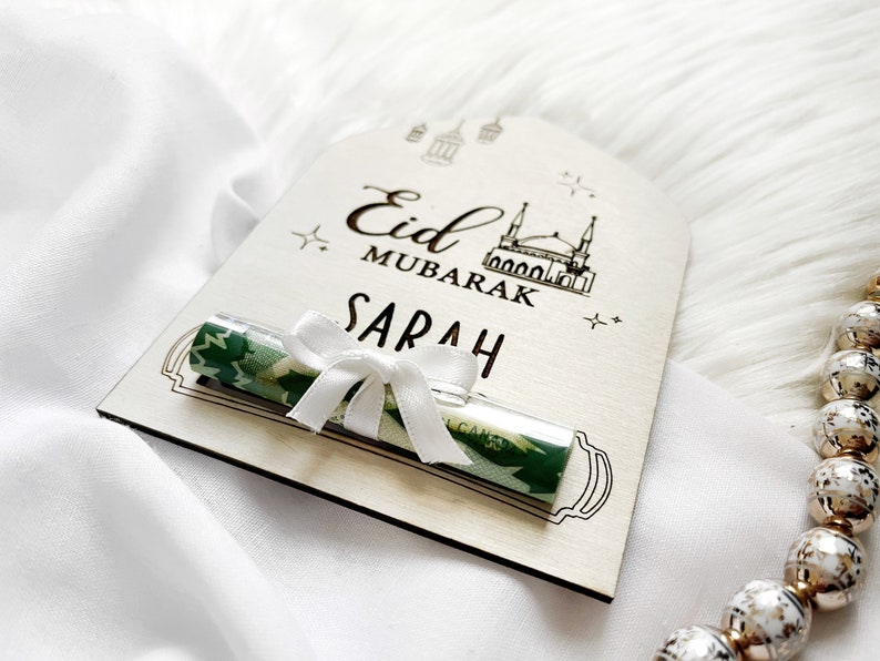 Puede incluir: Una tarjeta de madera con el texto "Eid Mubarak Sarah" y una tarjeta de regalo enrollada verde y blanca con una cinta blanca. La tarjeta tiene un dibujo de una mezquita y estrellas.