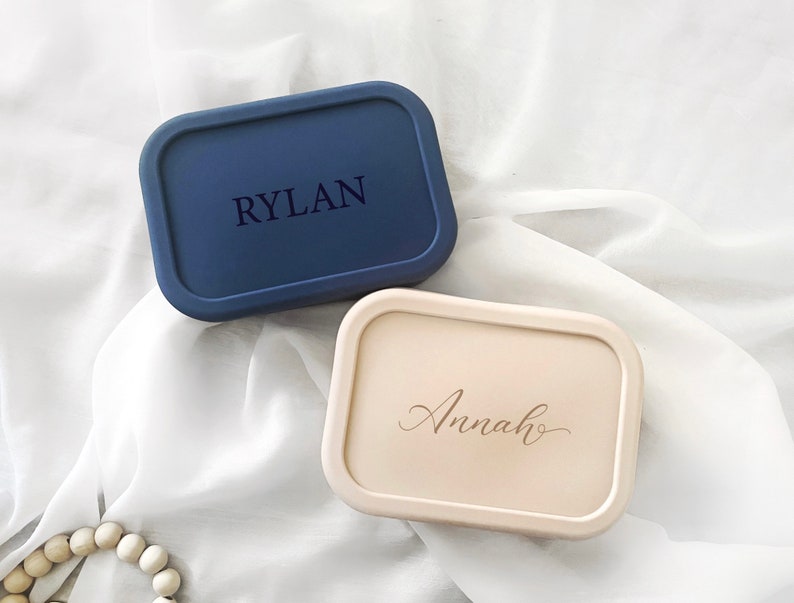 Pode incluir: Duas caixas retangulares personalizadas. Uma &eacute; azul marinho com o nome "RYLAN" gravado na tampa. A outra &eacute; bege claro com o nome "Annah" gravado na tampa. Ambas as caixas t&ecirc;m bordas arredondadas.