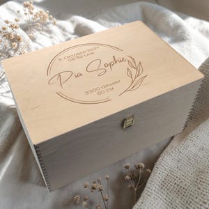 Peut inclure: Coffret souvenir en bois clair avec couvercle à charnière et fermoir en métal. Le couvercle est gravé du nom "Pia Sophie", de la date de naissance, de l'heure, du poids (3300 grammes) et de la taille (50 cm). Un motif floral est également gravé.