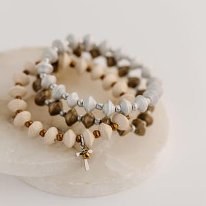 May include: Three beaded bracelets in neutral tones. The bracelets feature paper beads in cream, silver, and brown, strung together with small gold-colored beads. One bracelet has a small silver cross charm with a gold accent.