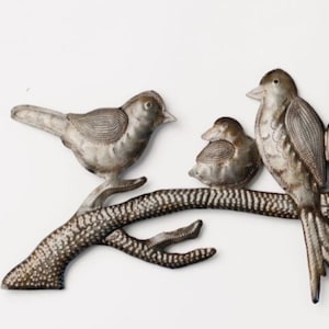 May include: Metal wall art featuring three birds perched on a branch. The birds are silver with detailed feather patterns. The branch is brown with white speckles, set against a white background. A decorative piece for home decor.
