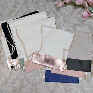 Könnte beinhalten: Eine Sammlung von Turnbeuteln in verschiedenen Farben, darunter Weiß, Schwarz und Grau. Jede Tasche hat eine kontrastierende Unterseite in Metallic-Roségold, Rosa oder Blau. Die Taschen haben beigefarbene Kordelzüge und sind auf einer gemusterten Oberfläche angeordnet.