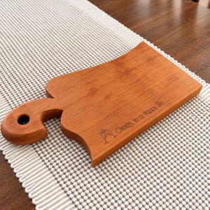 May include: A rectangular wooden serving board with a curved handle featuring a circular hole. The board has a smooth, light brown finish and is engraved with the text Cheers to a Happy Life and an icon of two clinking wine glasses.