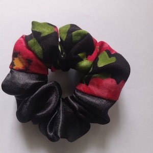 Puede incluir: Un scrunchie negro con estampado floral rojo y verde. El scrunchie está hecho de satén y tiene un diseño fruncido.