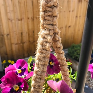 May include: A close-up of a macrame plant hanger made with natural jute rope. The hanger is attached to a metal ring and is holding a pot of purple pansies.