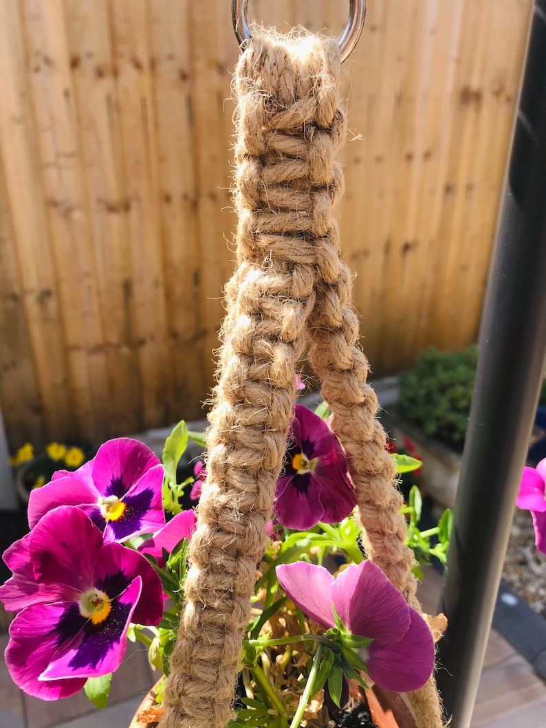 May include: A close-up of a macrame plant hanger made with natural jute rope. The hanger is attached to a metal ring and is holding a pot of purple pansies.
