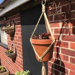 May include: A terracotta pot with a green plant hanging from a macrame plant hanger attached to a metal bracket on a brick wall.