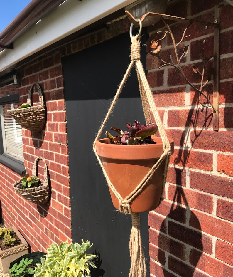 May include: A terracotta pot with a green plant hanging from a macrame plant hanger attached to a metal bracket on a brick wall.